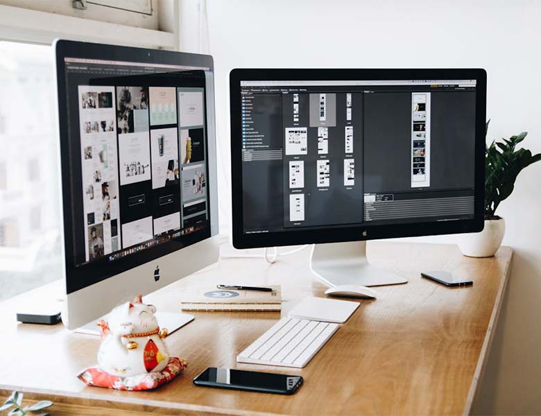 A workspace setup with two monitors displaying design projects a keyboard mouse smartphone notebook and a decorative cat figurine on a wooden desk