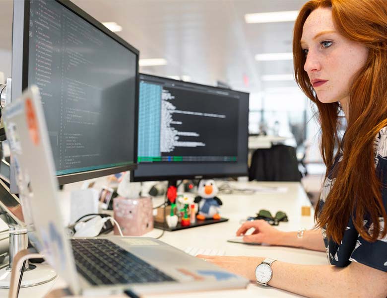 A person with long hair works at a desk with multiple monitors displaying code typing on a keyboard in an office setting