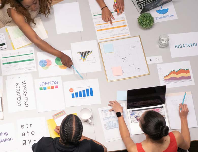 A diverse group of people work together at a table covered with charts graphs documents a laptop and office supplies analyzing data and discussing marketing strategies
