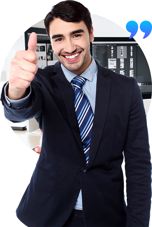 Man in a suit and tie giving a thumbs up and smiling standing in front of a blurred office background with a computer and paperwork